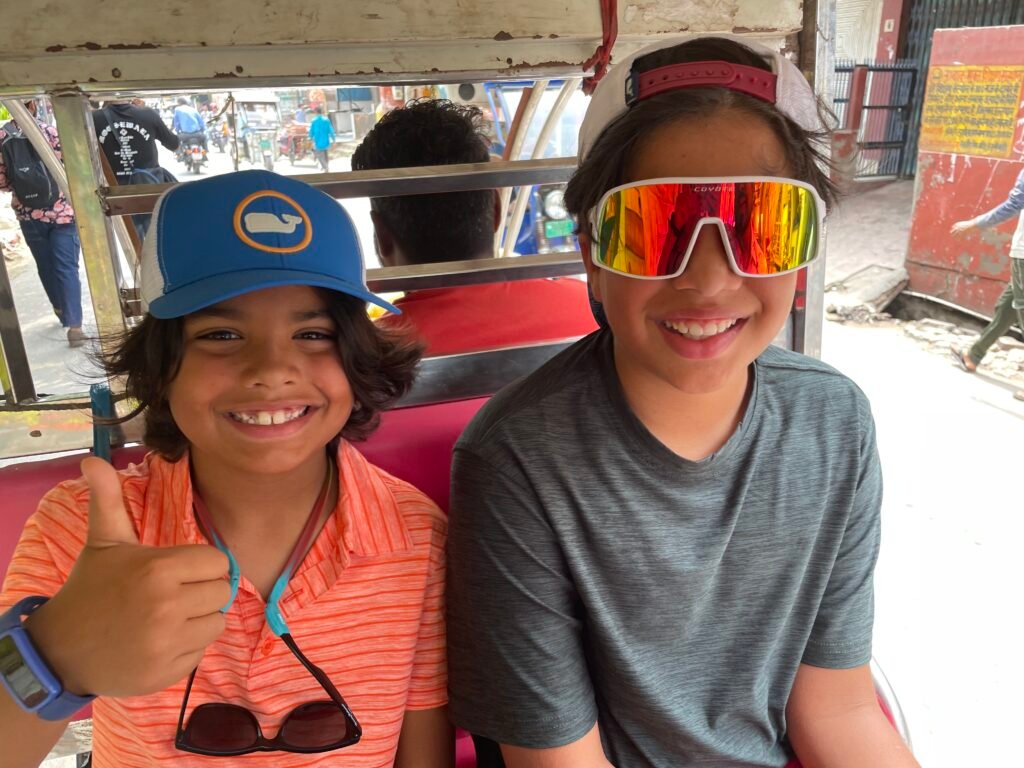 Caleb and Micah smiling and giving a thumbs up while riding in an auto rickshaw on a busy street in India