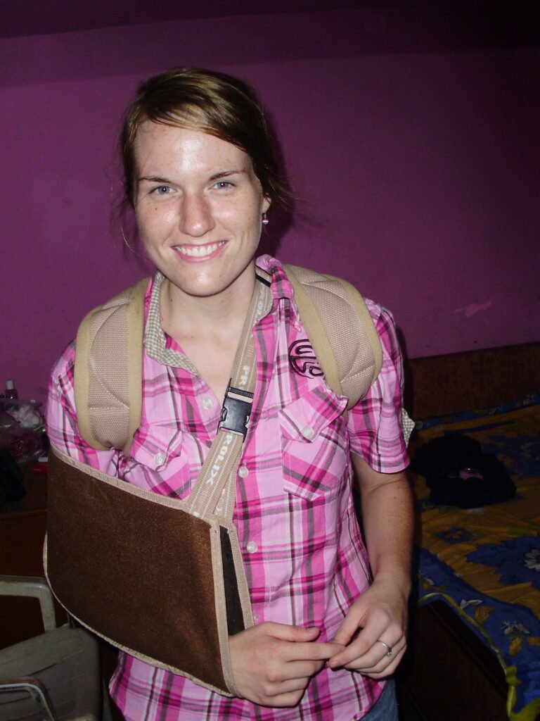 Kelly Damor in her early twenties in India, wearing an arm sling after breaking her scapula, smiling in a guest house room